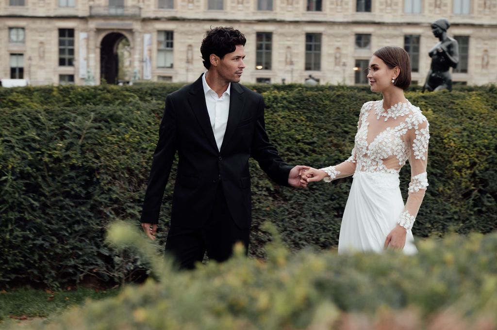 Elegant couple in formal attire holding hands in a lush garden, woman in lace bridal gown, man in black suit.