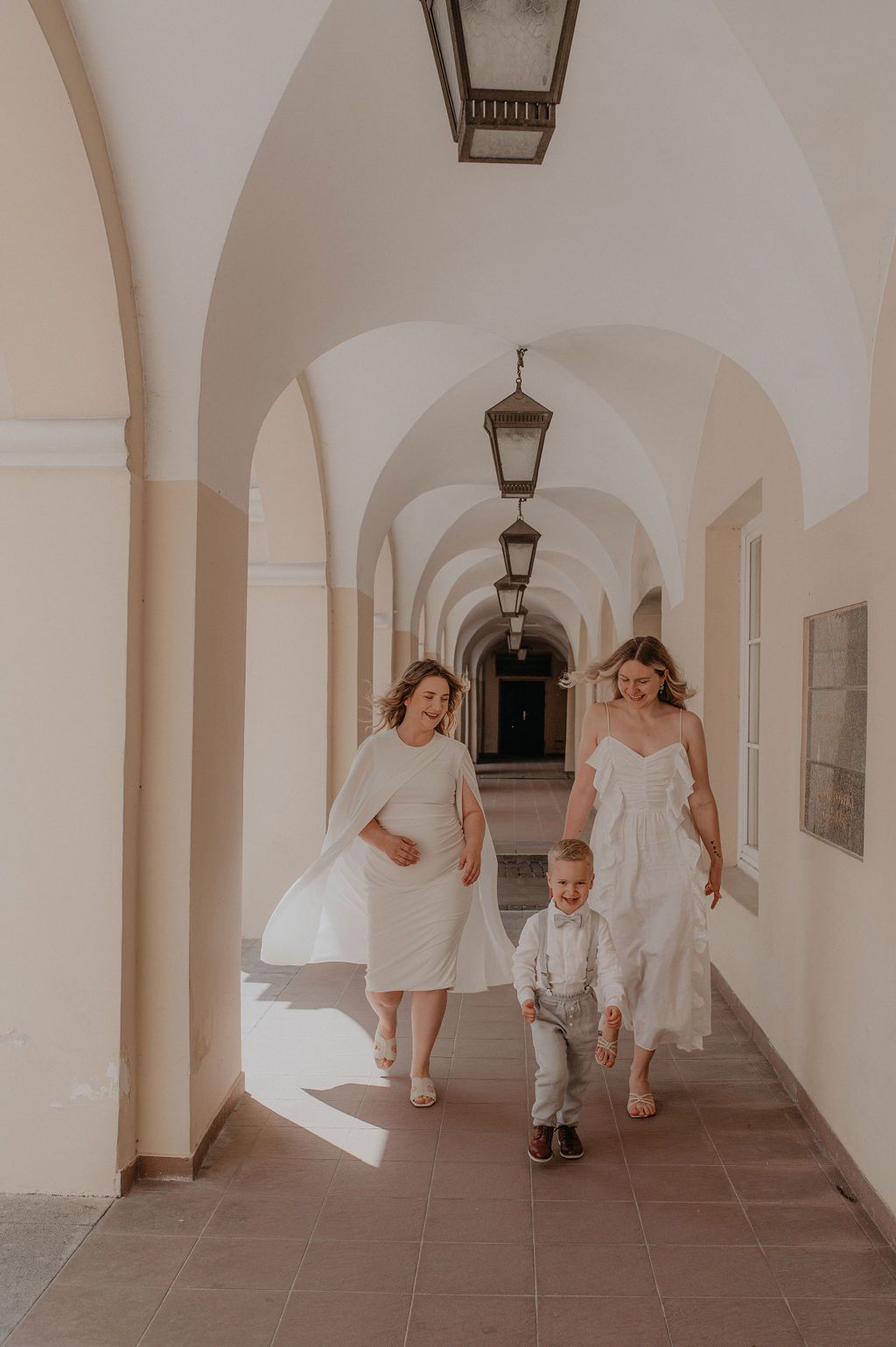 Two women in white dresses walk with a toddler boy through an elegant arched corridor with hanging lanterns.