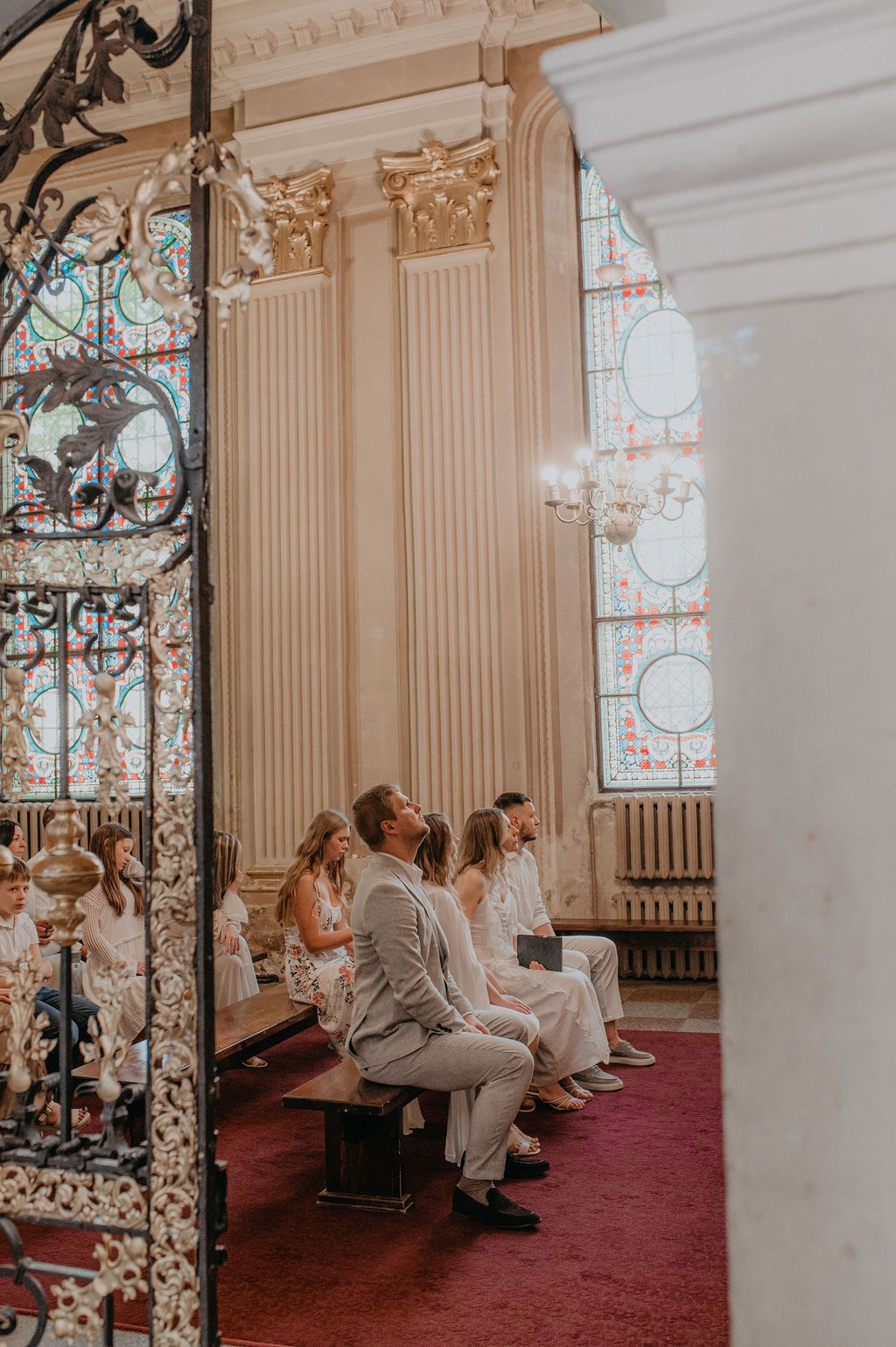 Guests seated in elegant church pews beneath stained glass windows during a wedding ceremony.