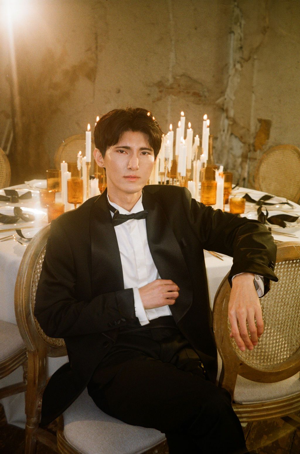 Groom in black tuxedo sitting by candlelit reception table with gold chairs in moody venue.