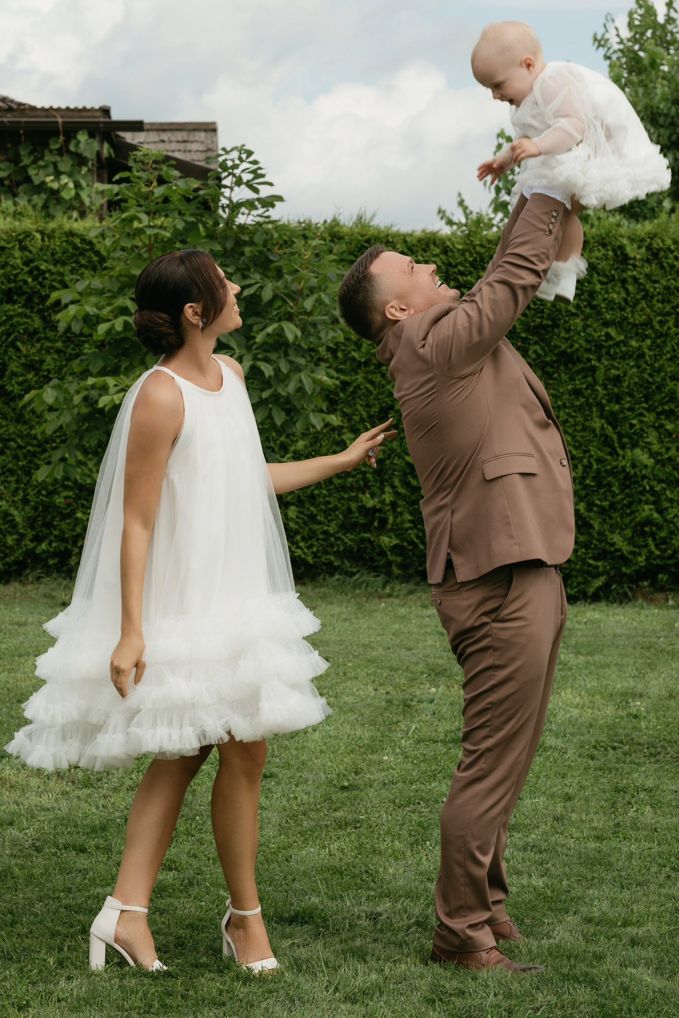 Bride in white ruffled dress and groom in brown suit lifting baby in garden on wedding day.