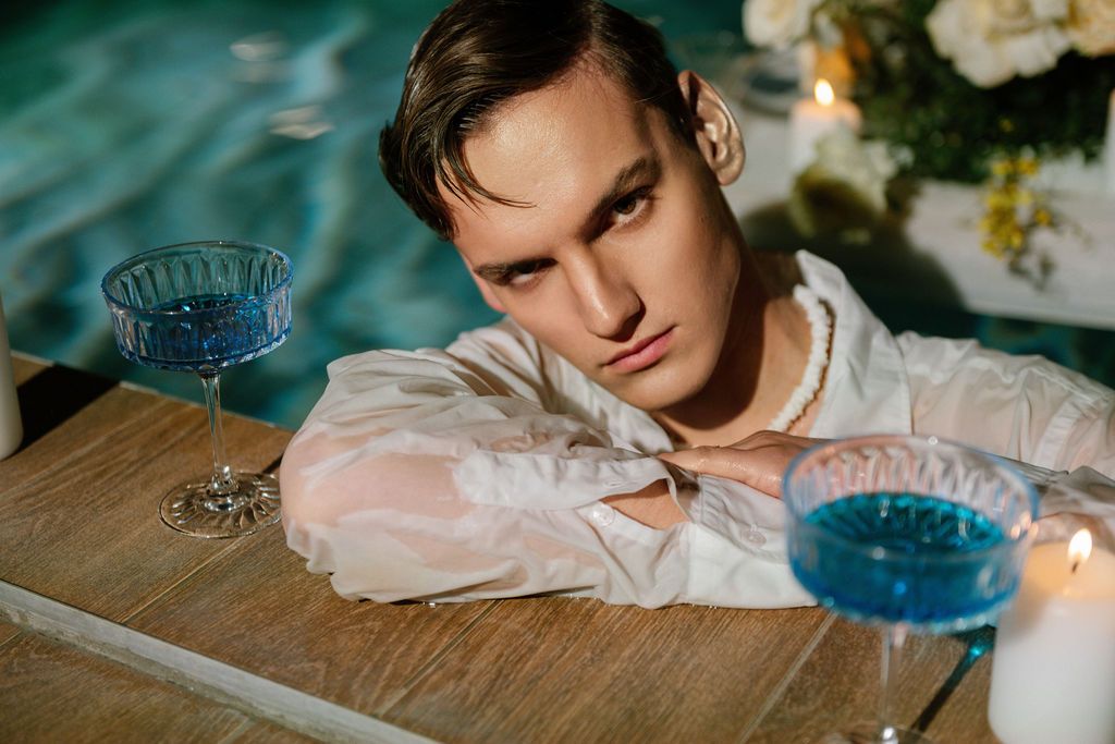 Young man in a white sheer shirt and pearl necklace rests his arms on a wooden table beside blue cocktails and candles near a pool.