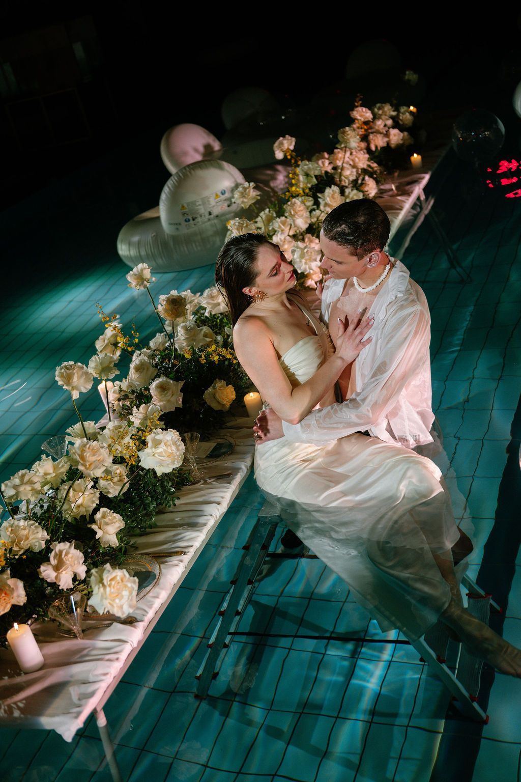 Couple in white embracing on submerged steps in a candlelit pool surrounded by cream roses and floral arrangements at night.