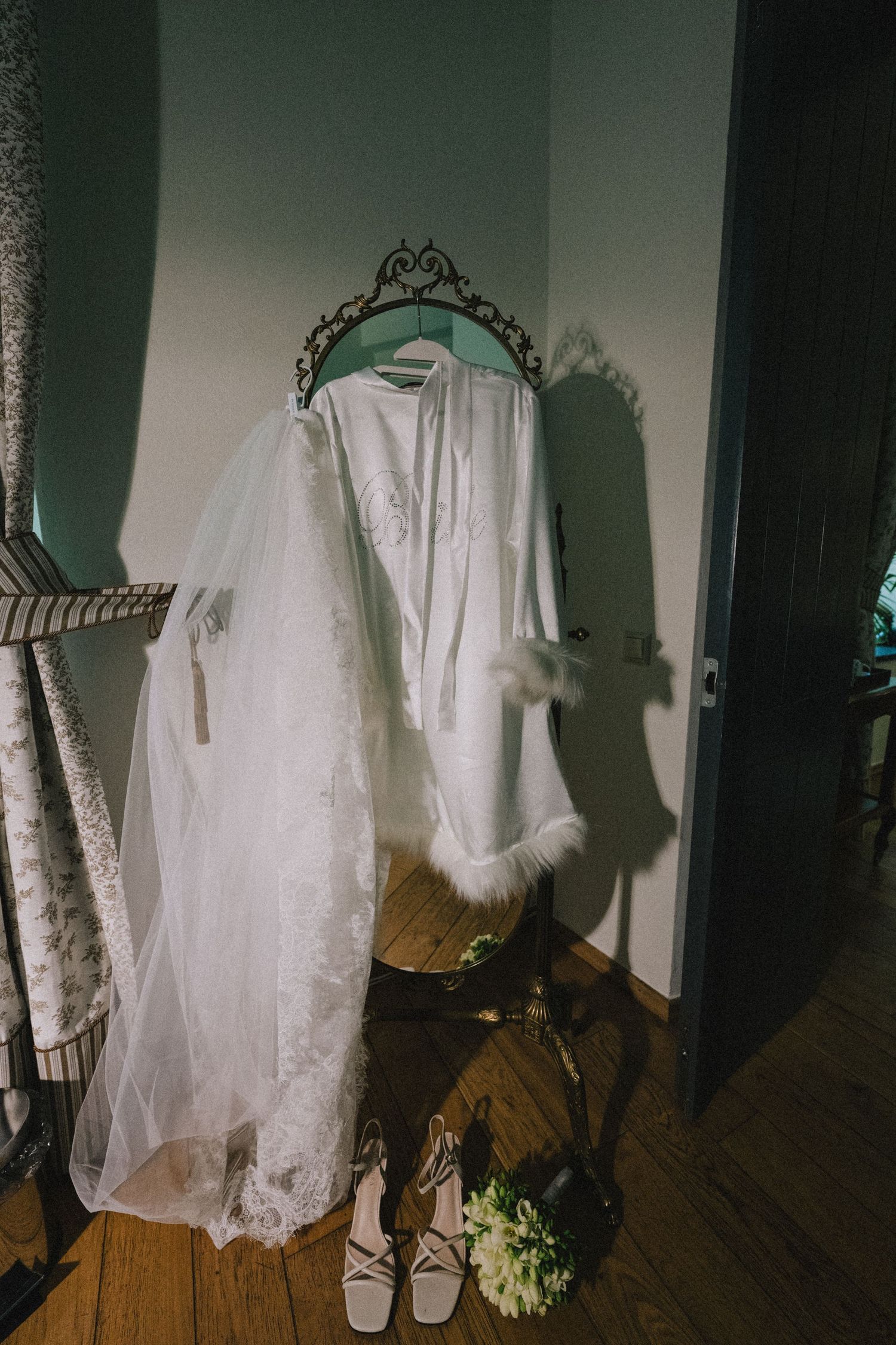 White bridal robe and lace veil hanging on an ornate stand beside bridal shoes and a white floral bouquet on a wooden floor.