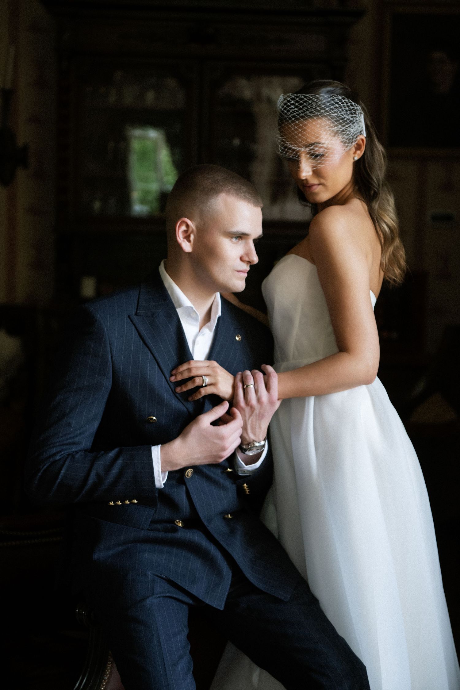 Bride in a white strapless gown and birdcage veil poses beside a groom in a navy pinstripe suit in a dimly lit, elegant room.