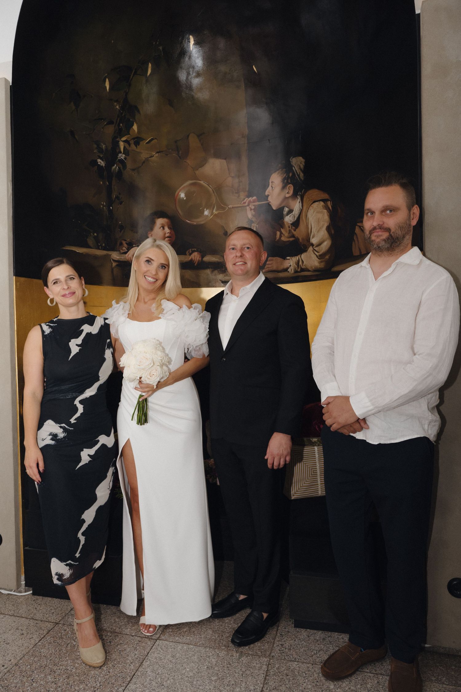A bride in a white slit gown holding a bouquet poses with a groom and two guests in front of a large dark Baroque painting.