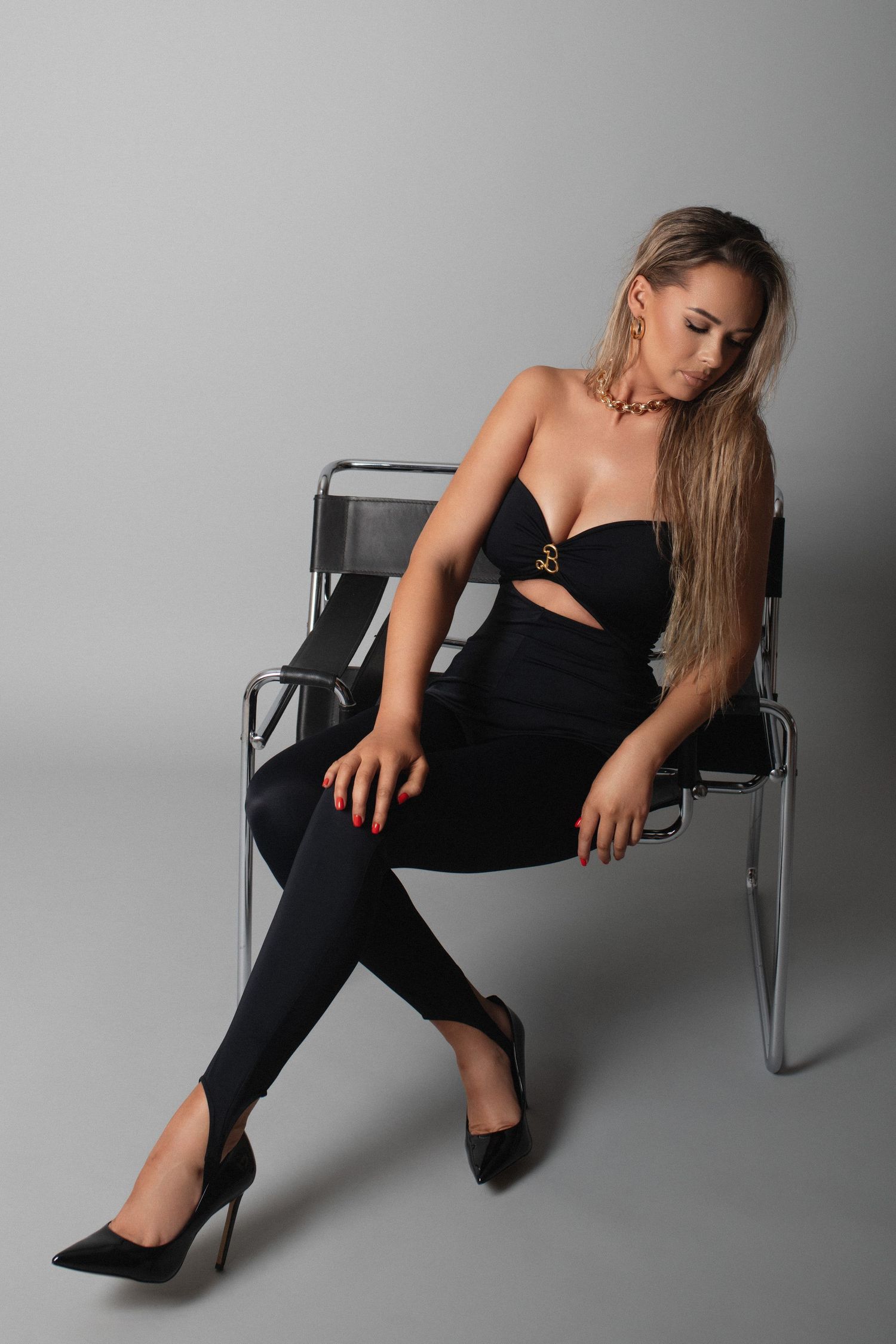 Woman in black strapless jumpsuit and heels sitting on chrome chair against gray background.