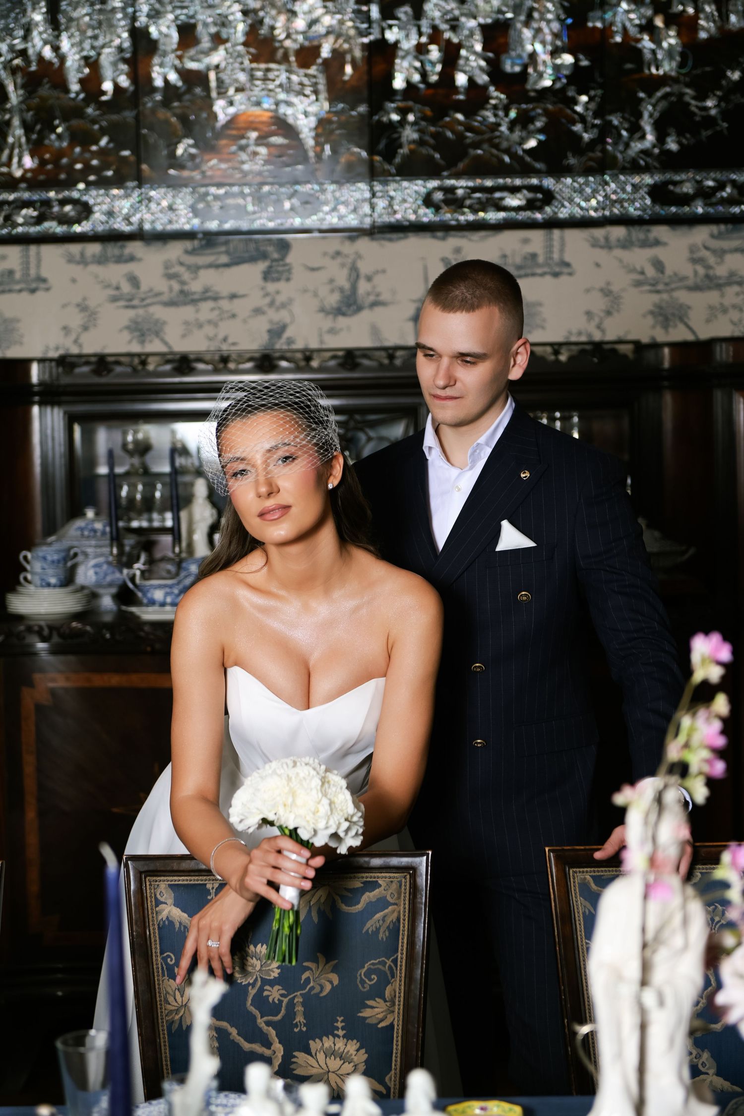 Bride in a strapless white gown and birdcage veil holding a bouquet beside a groom in a pinstripe suit in an elegant, ornate dining room.