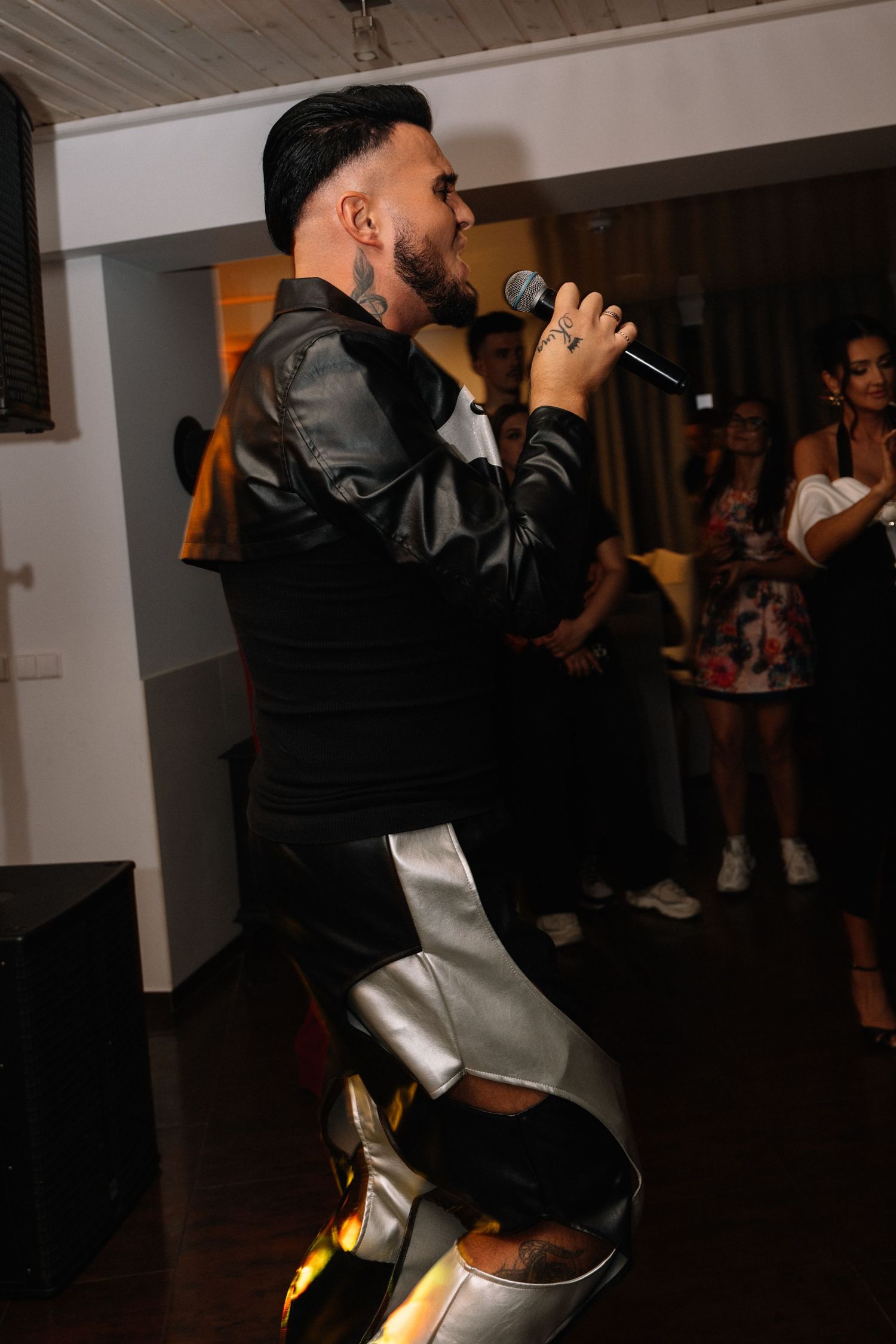 A man in a black and silver leather outfit sings into a microphone at an indoor party while guests watch in the background.