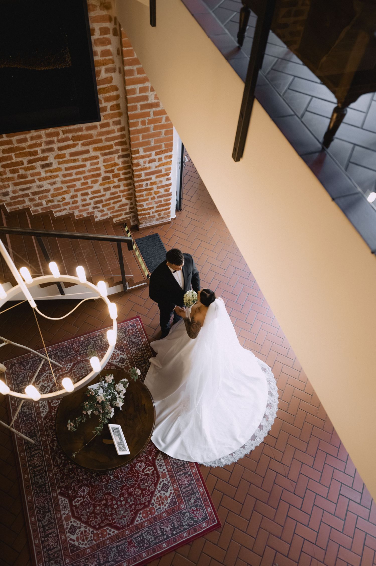 Overhead view of a bride in a lace-trimmed gown and a groom in a dark suit standing on a herringbone brick floor near a round table.
