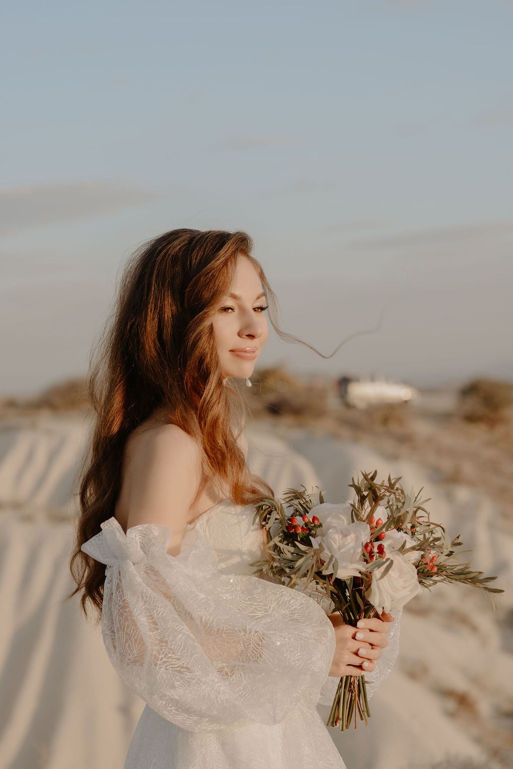 Bride in off-shoulder lace gown holds wildflower bouquet on sandy beach at golden hour.