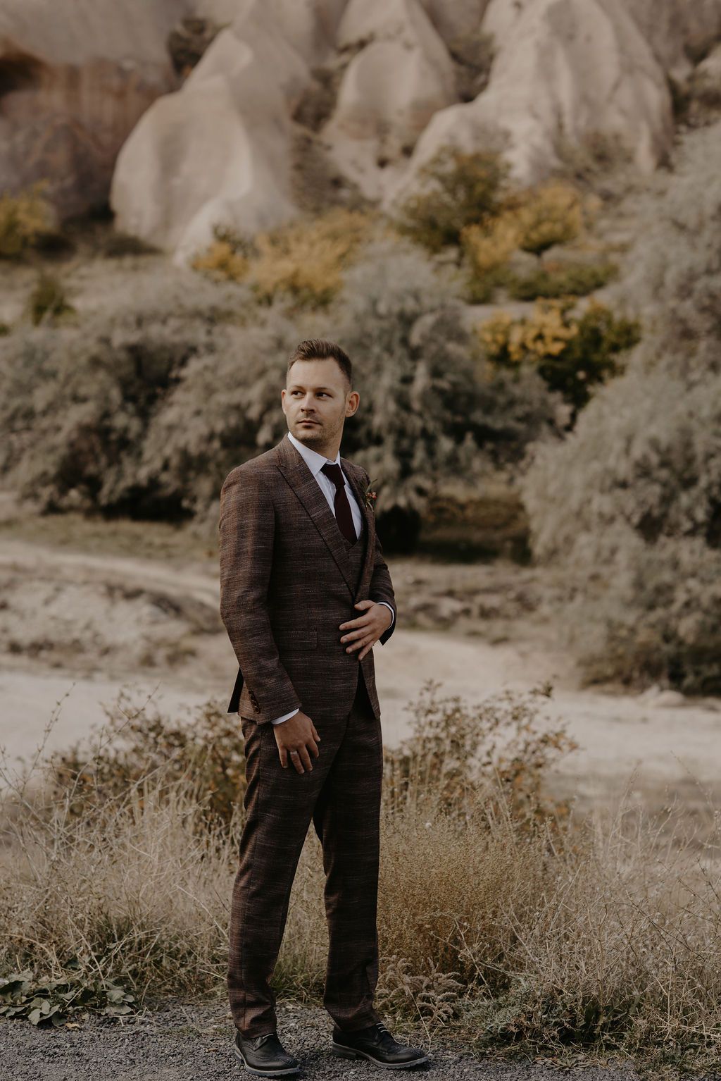 A well-dressed groom in a brown plaid suit stands confidently in a scenic rocky landscape with autumn foliage.