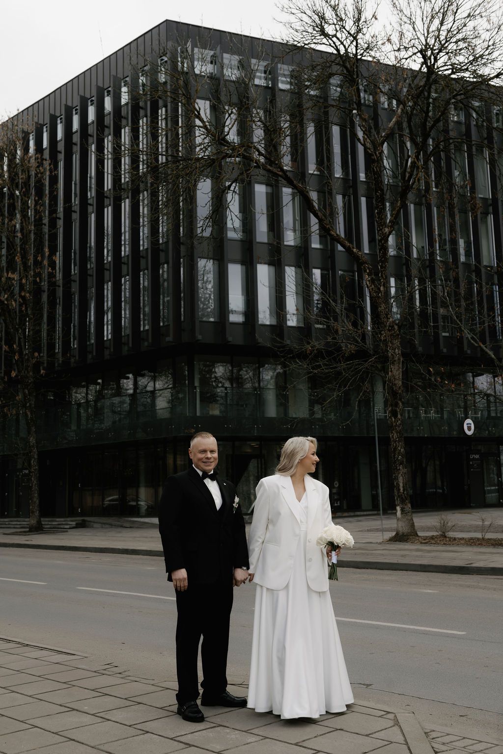 Elegant wedding couple in formal attire holding hands on a city street with a modern black building backdrop.