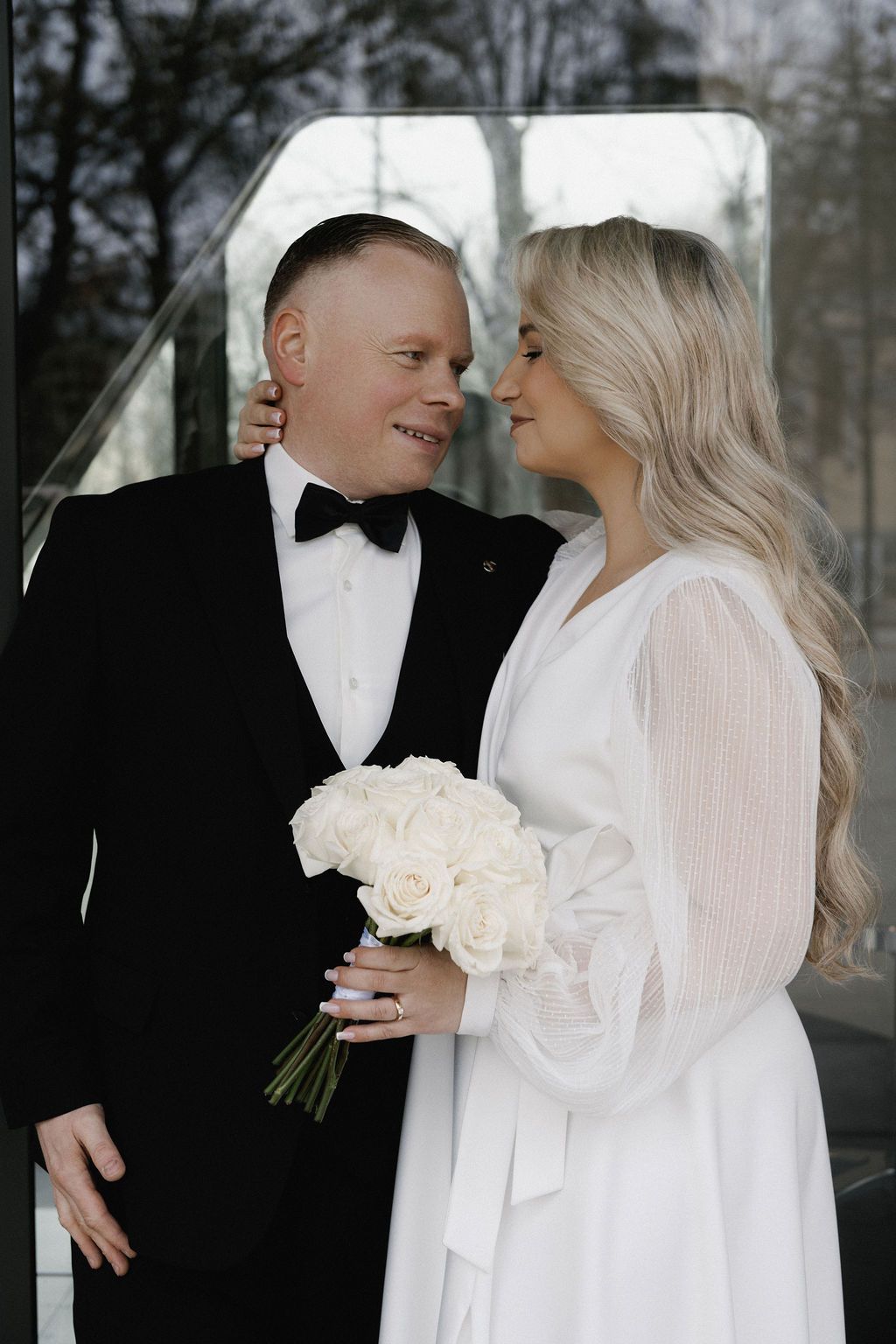 A bride and groom in elegant wedding attire share a romantic moment, she holds a white rose bouquet.