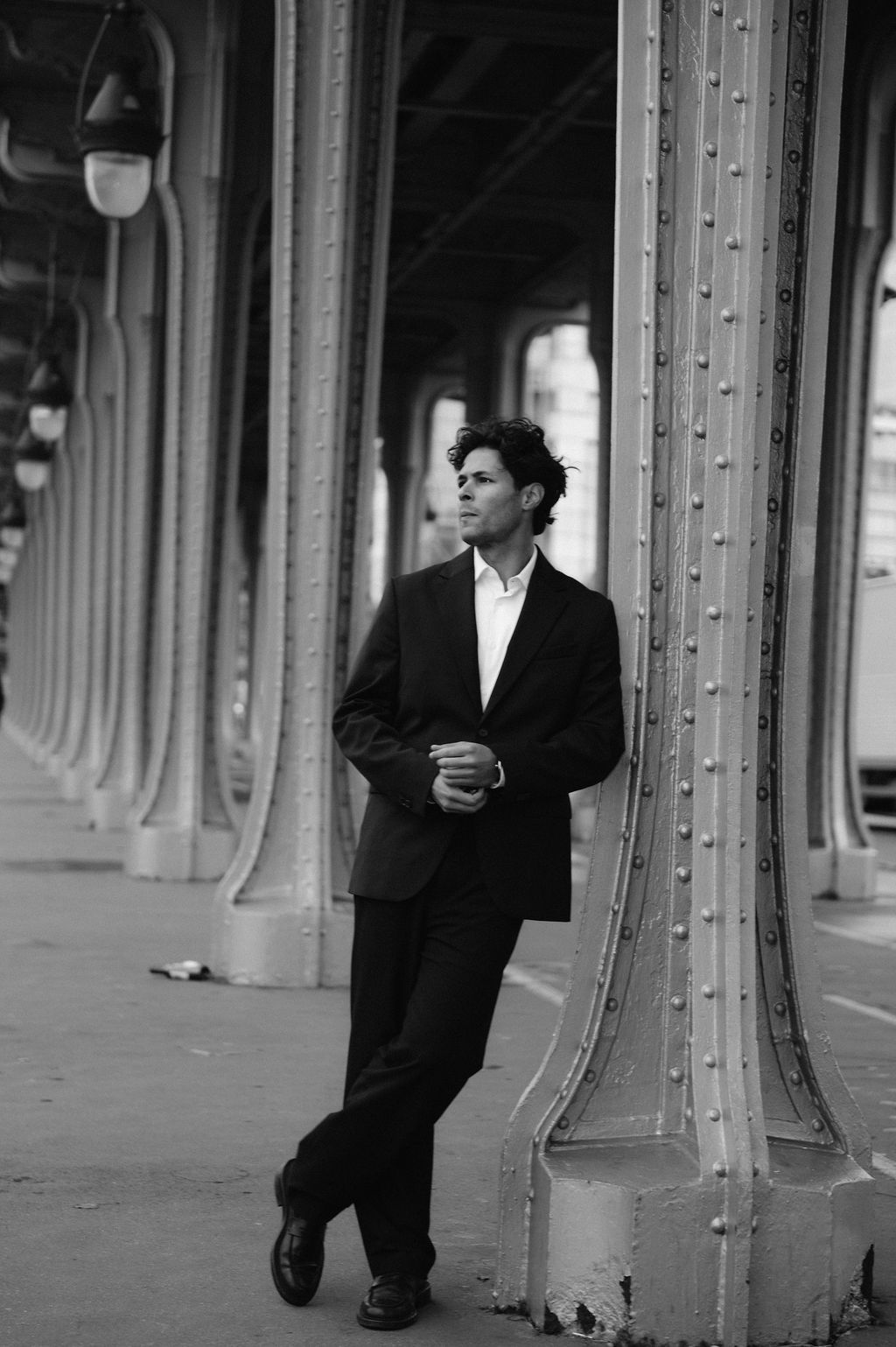 A man in a black suit leans against a ornate metal column in a black and white urban architectural setting.