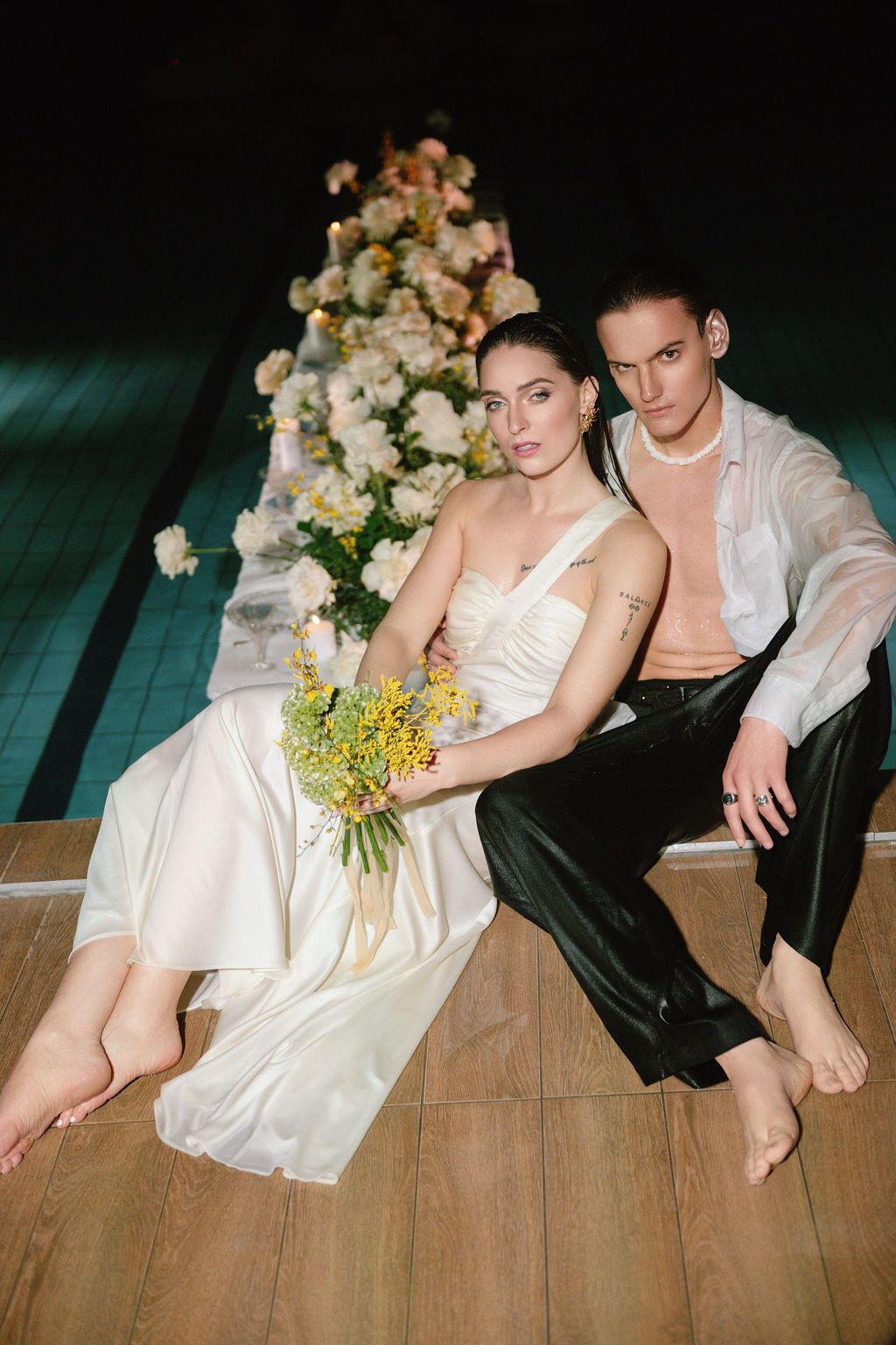 Bride in white satin gown and groom in black pants sit barefoot on floor beside elegant floral table arrangement.