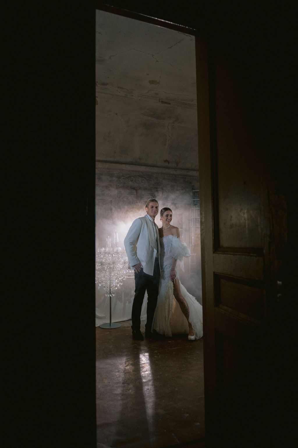 Couple in wedding attire framed by a dark doorway, bride in white gown and groom in suit, standing in softly lit room.