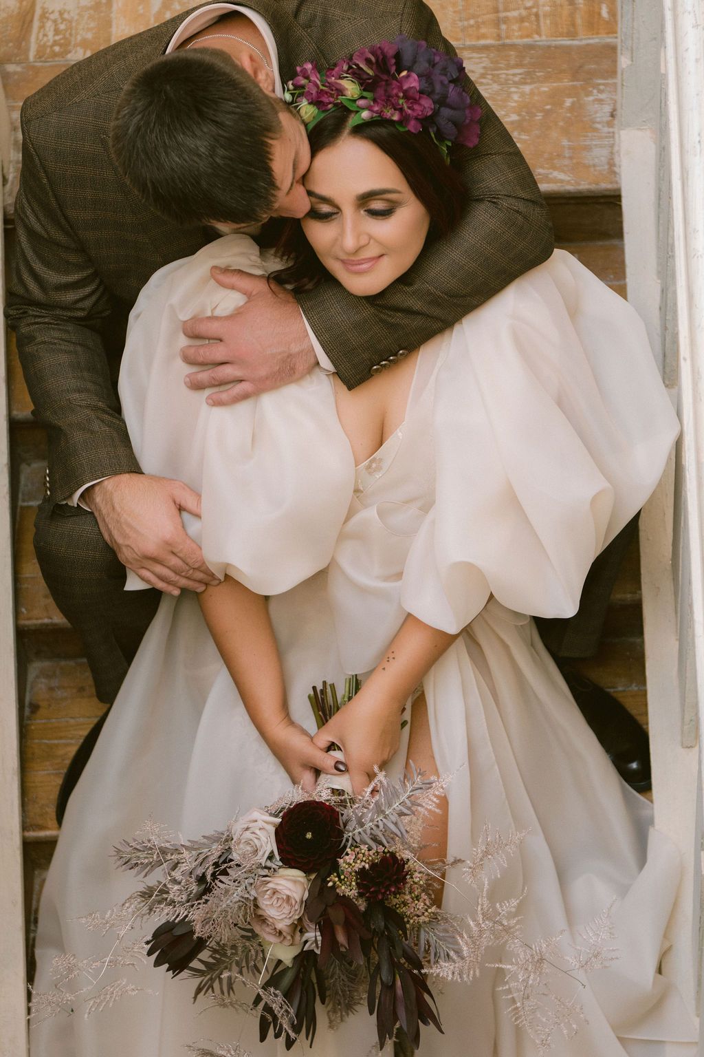 Bride in white gown with floral crown holds bouquet as groom embraces her tenderly on rustic wooden steps.