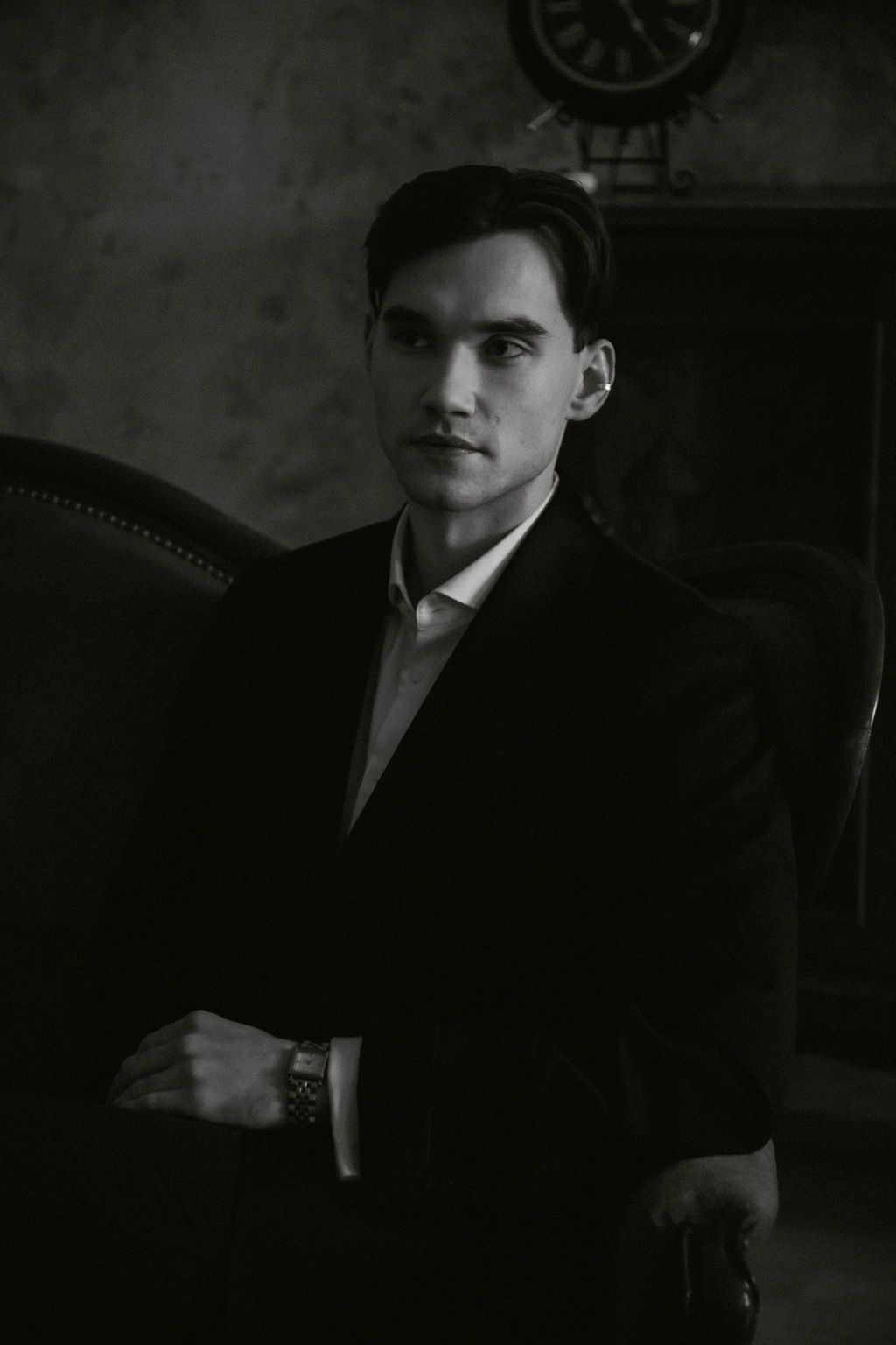 Black and white portrait of a young man in a suit, arms crossed, seated near a vintage clock in a dark room.