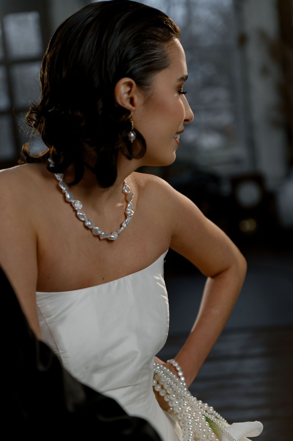 Elegant woman in strapless white gown wearing pearl necklace and earrings, hair swept up, viewed from behind.