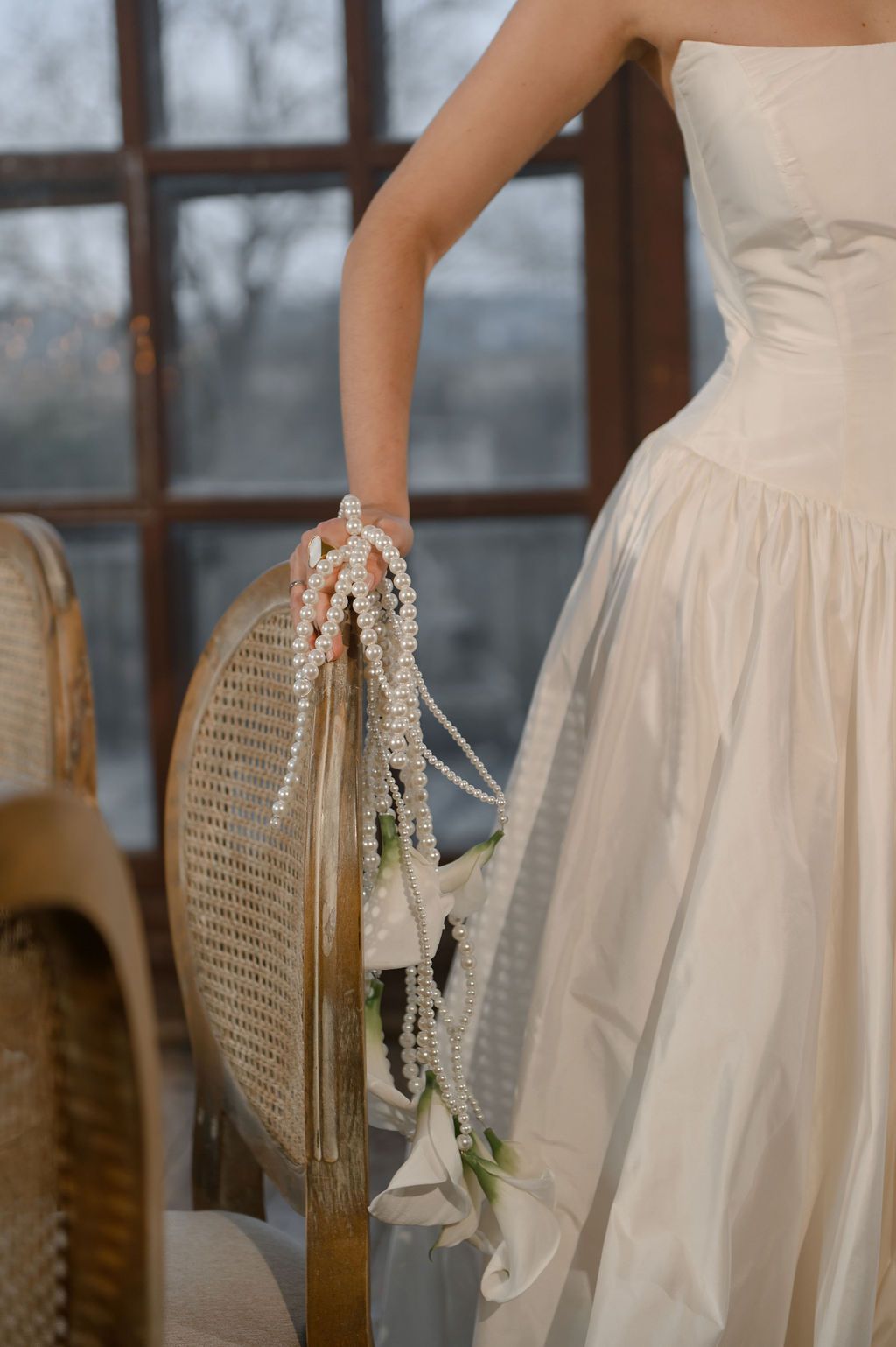 Bride in strapless ivory gown holding cascading pearl and floral bouquet beside a wicker chair near large windows.