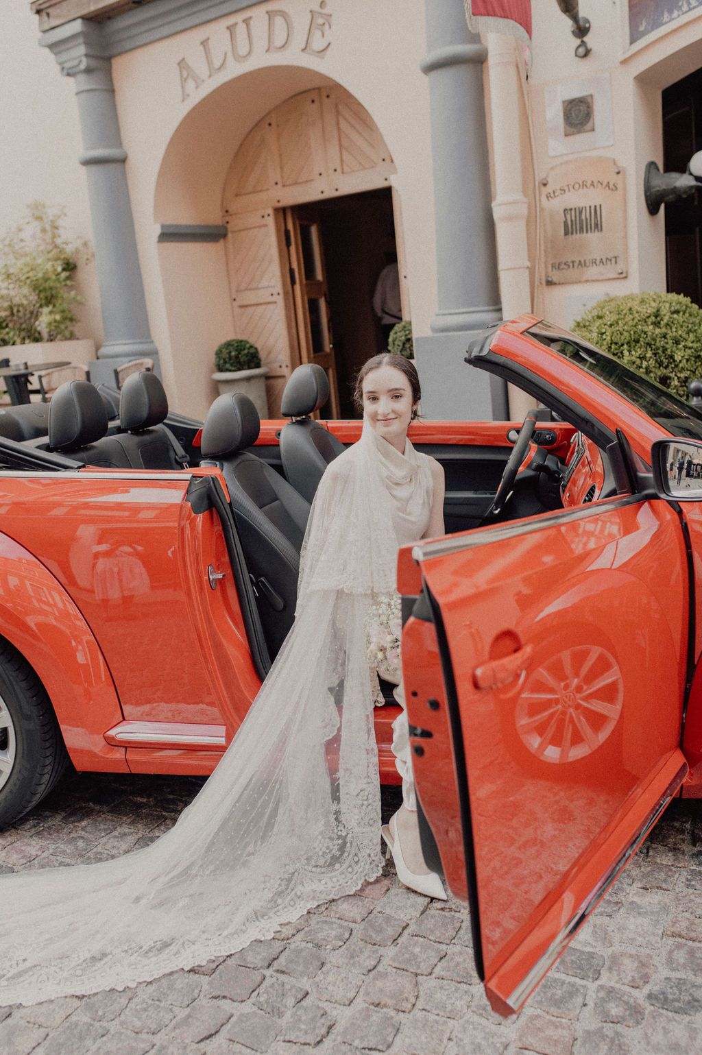 Bride in white lace gown and veil poses beside a red convertible outside the Alude building.