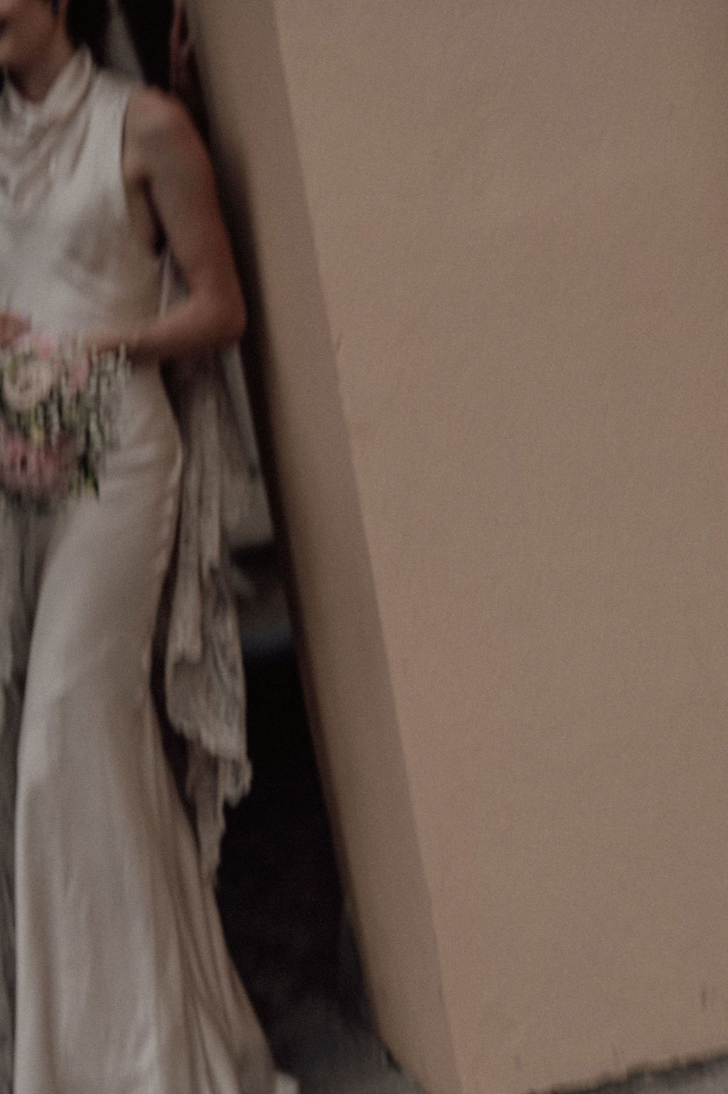 A bride in an elegant ivory sleeveless gown holds a floral bouquet while leaning against a beige wall.