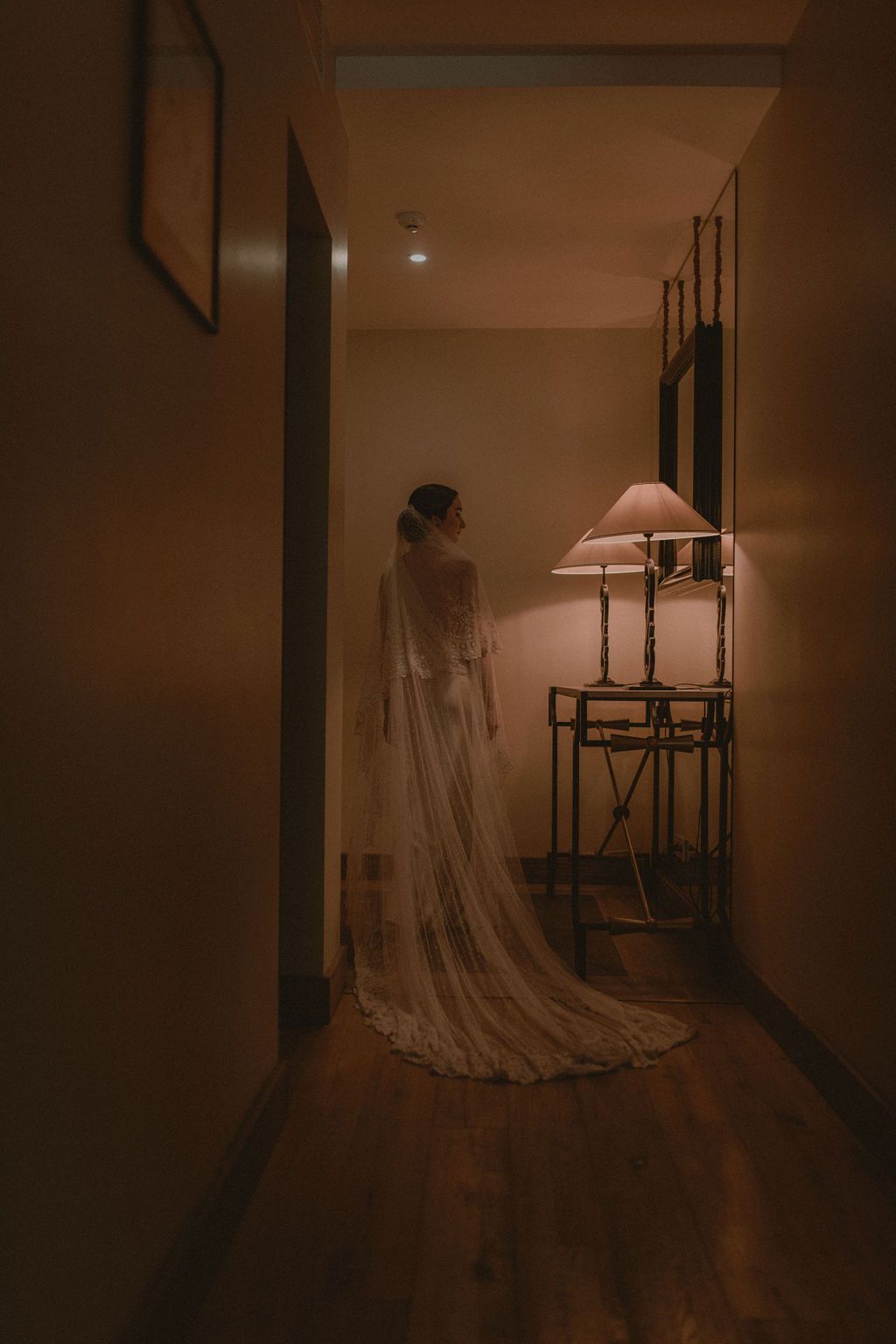 Bride in white wedding gown and long veil standing in dimly lit hotel hallway near glowing lamp.