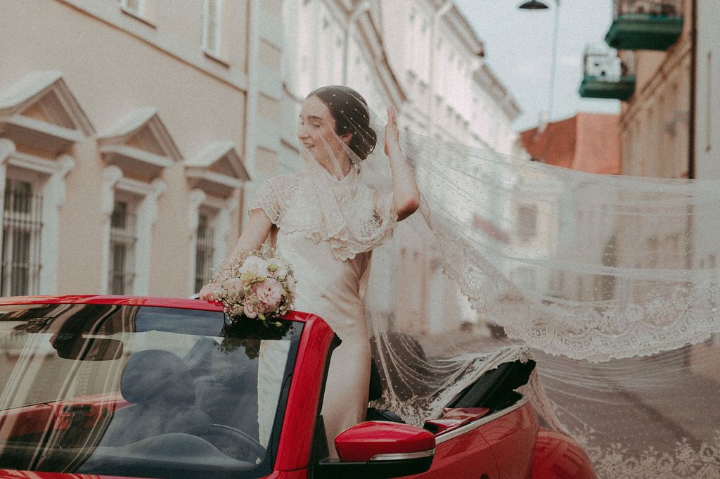 Bride in white wedding dress and veil sitting in red convertible car holding floral bouquet outdoors.