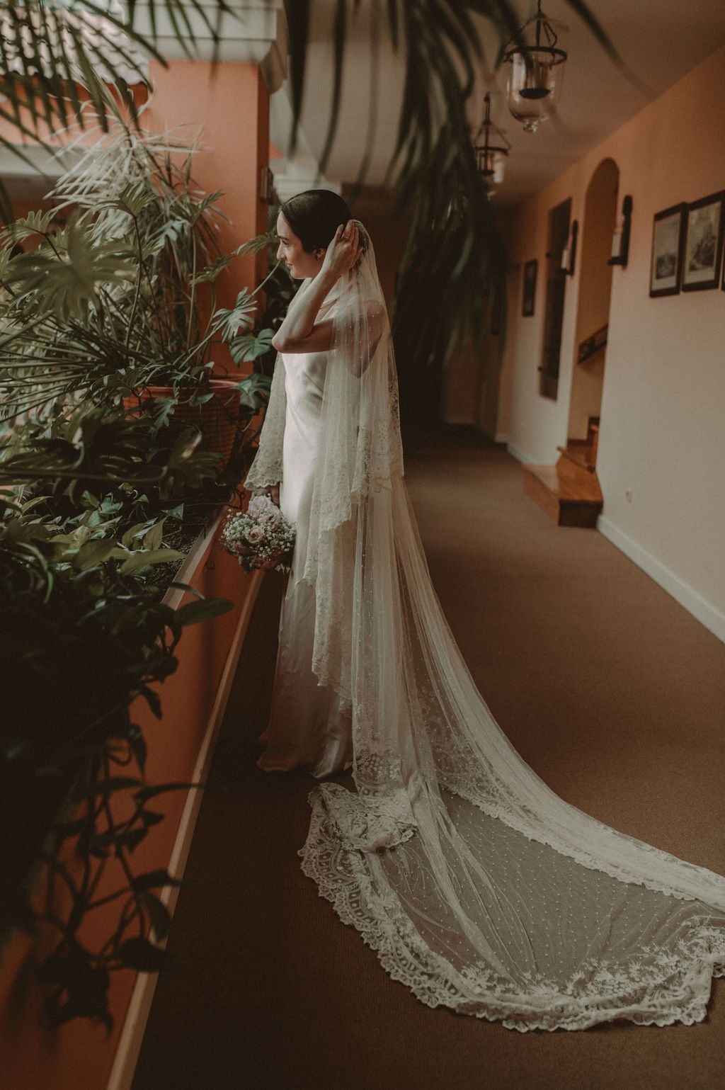 Bride in a stunning lace wedding gown with cathedral veil stands in an elegant hallway holding a bouquet.