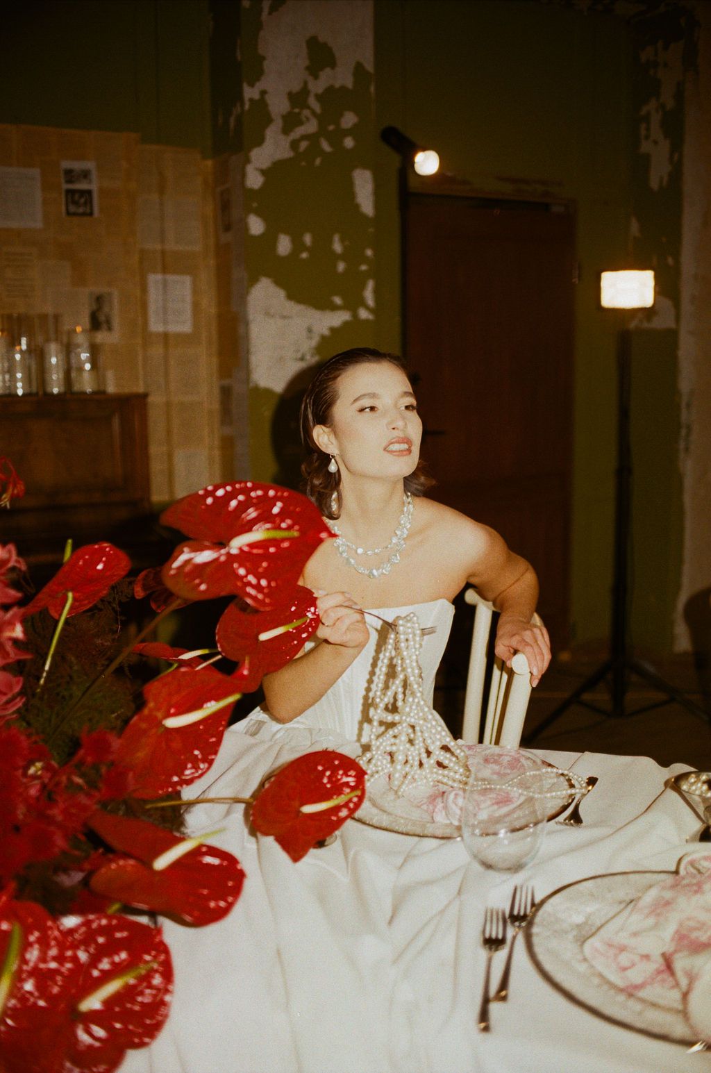 Elegant bride in white corset gown sits at wedding table adorned with red anthurium flowers and pearl jewelry.