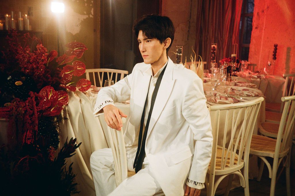 Stylish young man in white suit with black tie seated at elegant red-themed dinner table setting.
