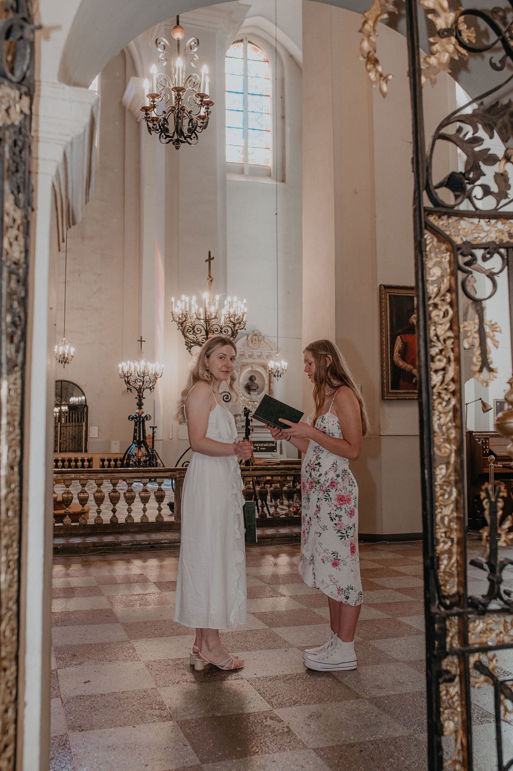 Two young women in dresses converse inside an elegant church with chandeliers and ornate iron gates.
