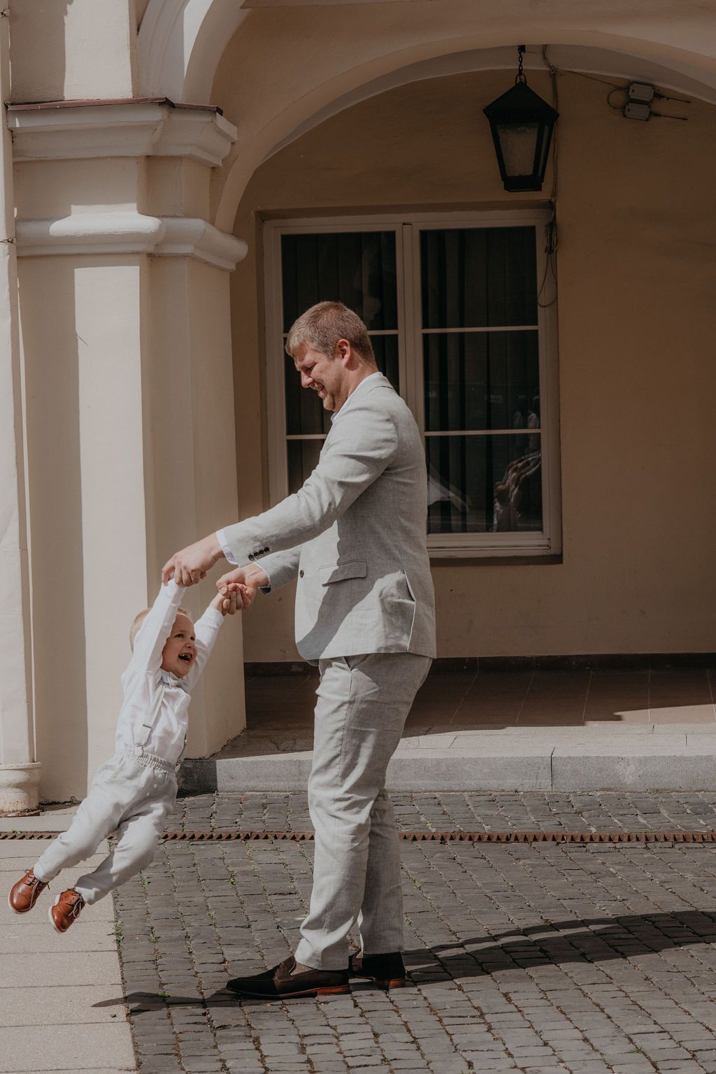A father in a light gray suit playfully swings his toddler son outdoors near an elegant building.