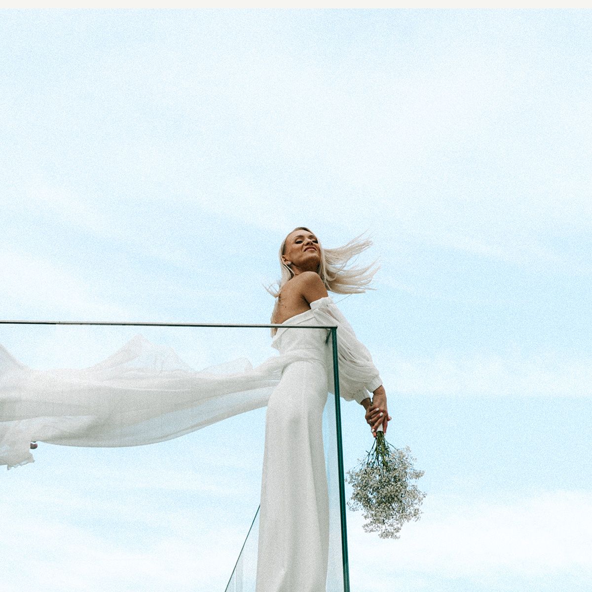 Bride in white gown holding bouquet on balcony, hair flowing in wind against blue sky.