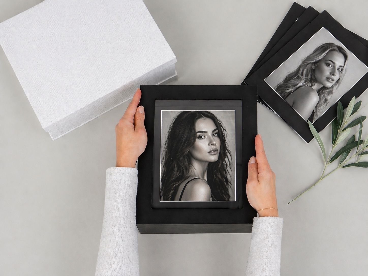 Hands holding a black framed portrait photo with additional framed prints and a white gift box on a light gray surface.
