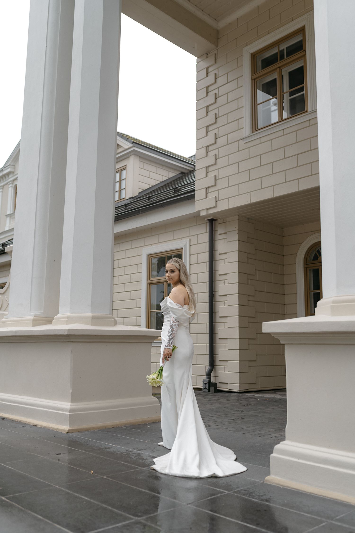 A bride in a white off-shoulder wedding gown poses elegantly between classic white columns in an architectural setting.