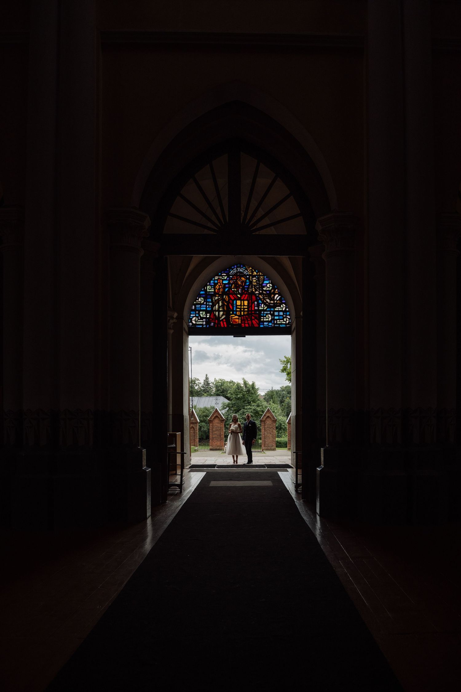 Couple silhouetted in ornate church doorway with stained glass arch, dark interior framing bright outdoor wedding scene.