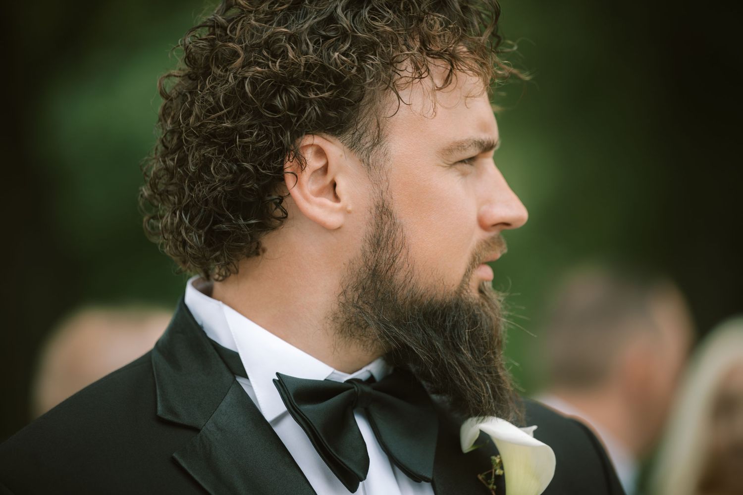 Bearded groom in black tuxedo with white boutonniere gazes solemnly during outdoor wedding ceremony.