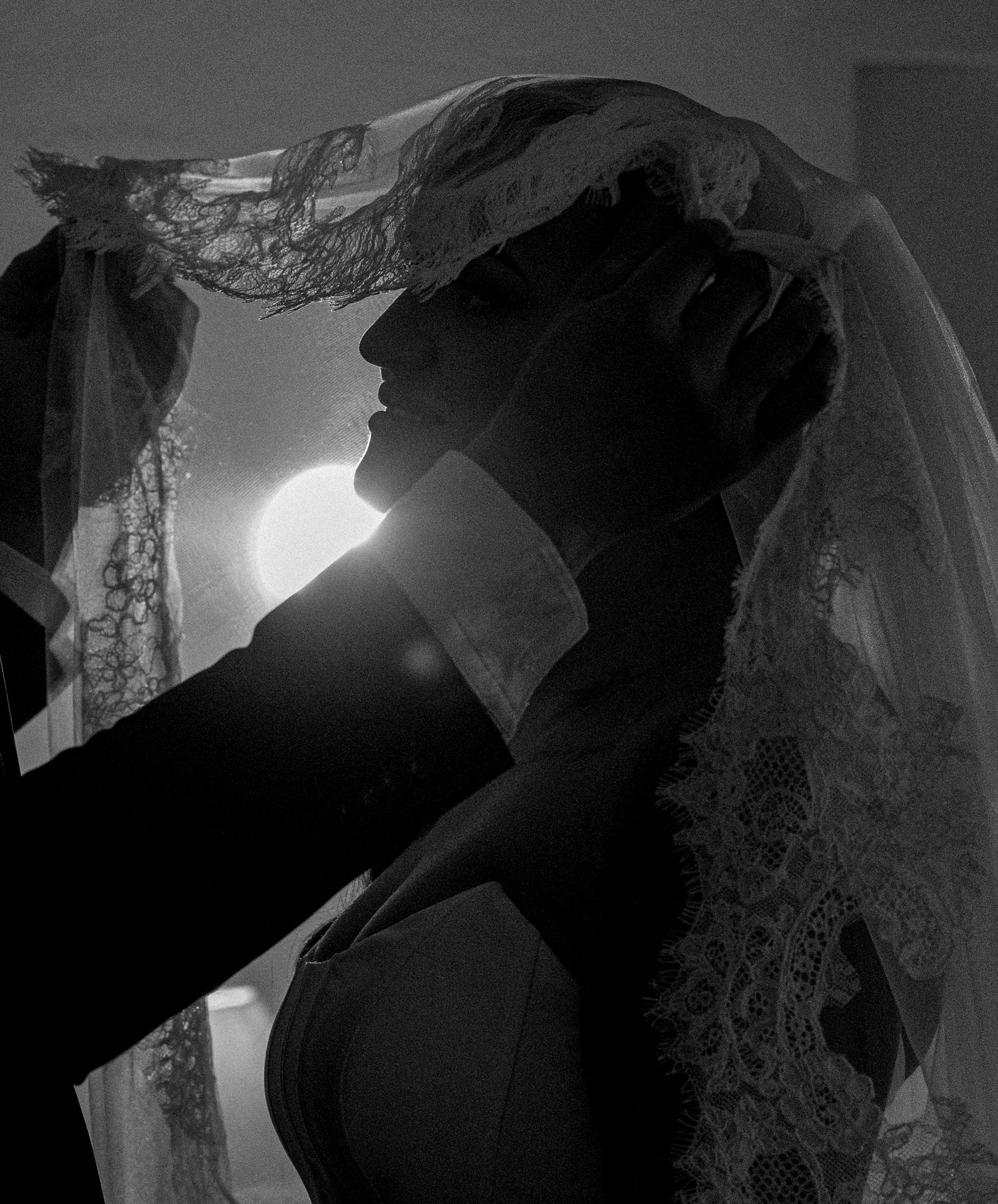 Black and white silhouette of a bride in a lace veil as a groom in a dark suit gently lifts it, with soft backlight glowing behind.