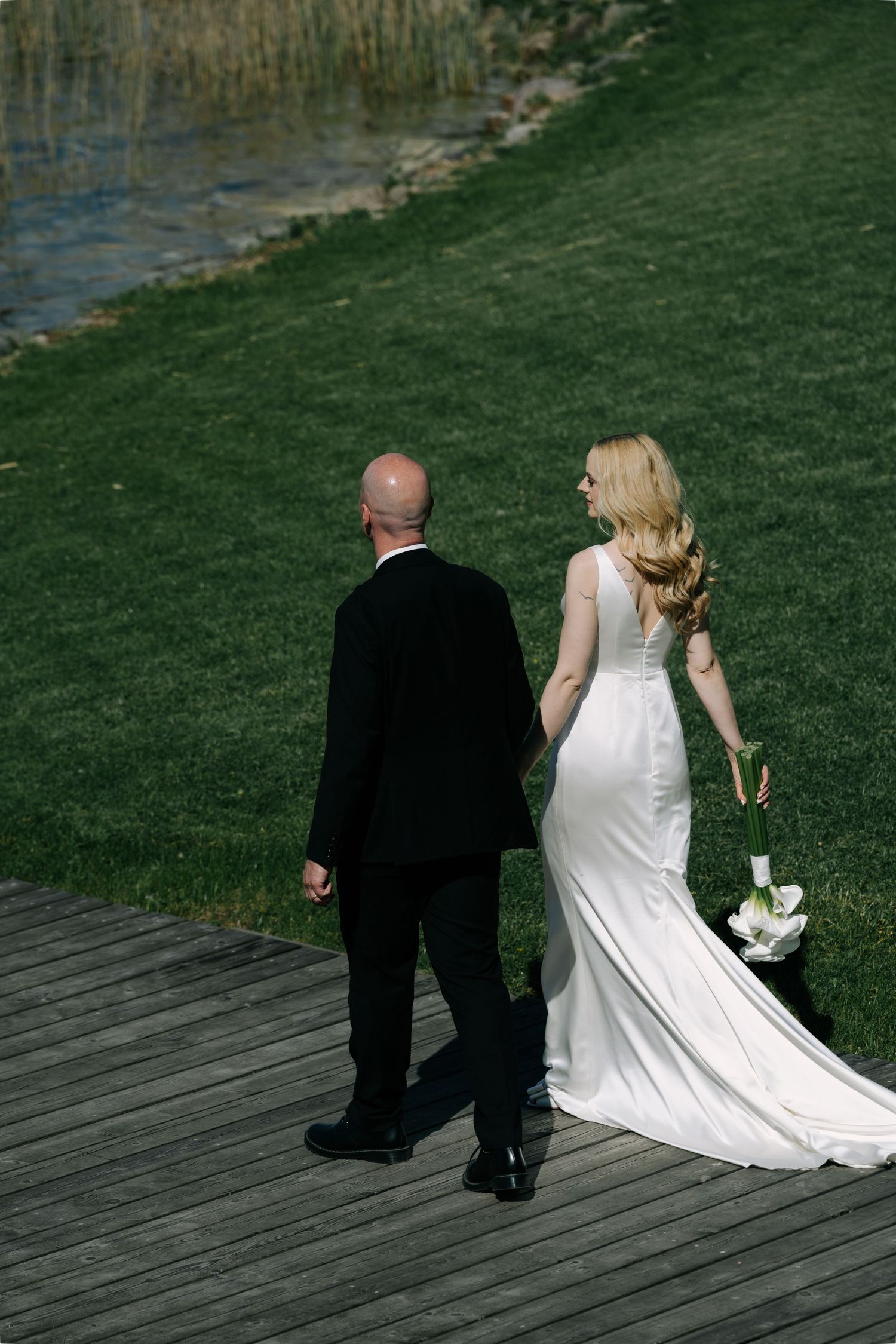 Bride in a white gown with a calla lily bouquet and groom in a black suit walk across a wooden dock beside a grassy lakeshore.