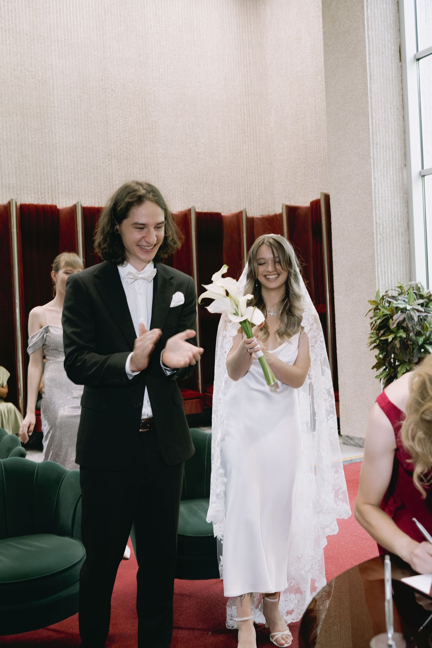 Smiling groom in a black tuxedo and bow tie applauds beside a bride in a white lace veil holding white calla lilies at a wedding ceremony.