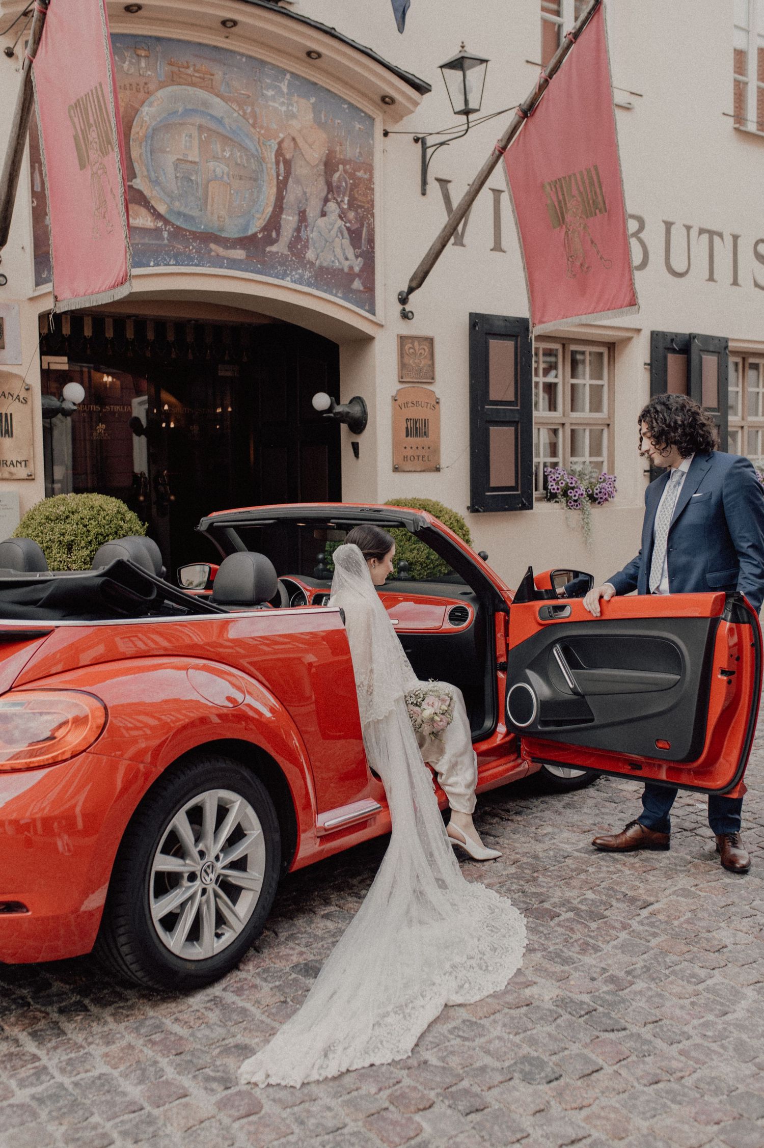 A bride in a white lace gown steps into an orange Volkswagen Beetle convertible on a cobblestone street.
