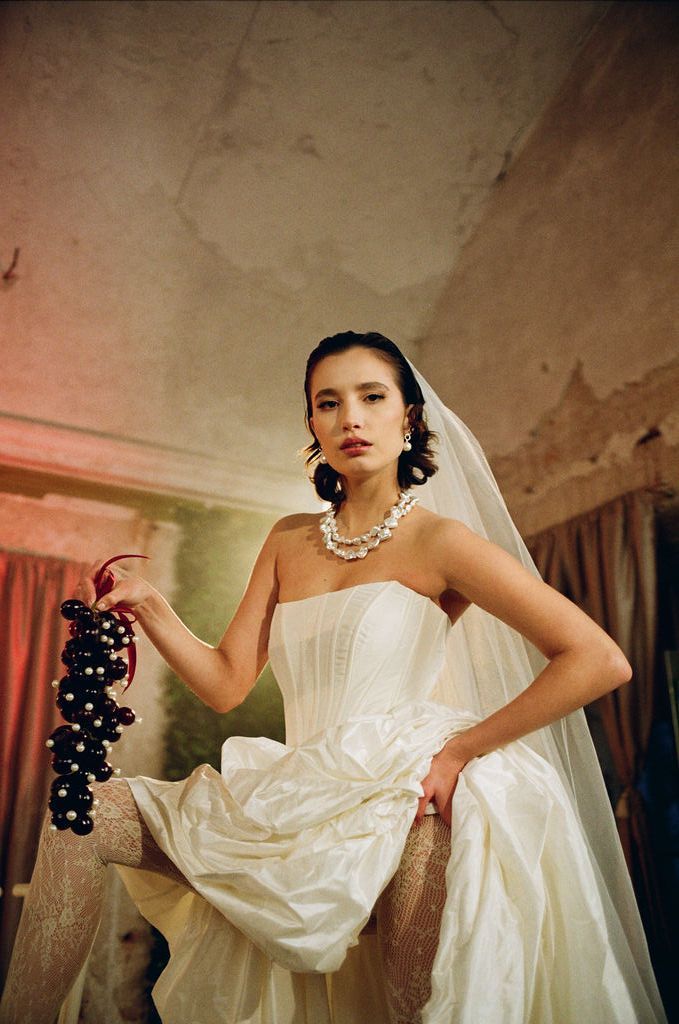 Bride in strapless white gown and veil holds dark grapes, wearing pearl necklace in vintage-styled editorial photo.