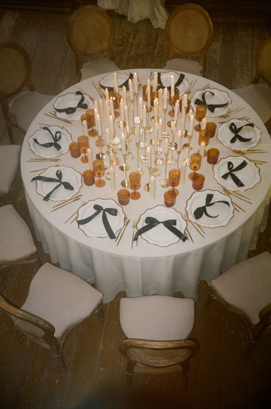 Elegant round wedding table with white linen, gold candles, and place settings adorned with dark green bow accents.