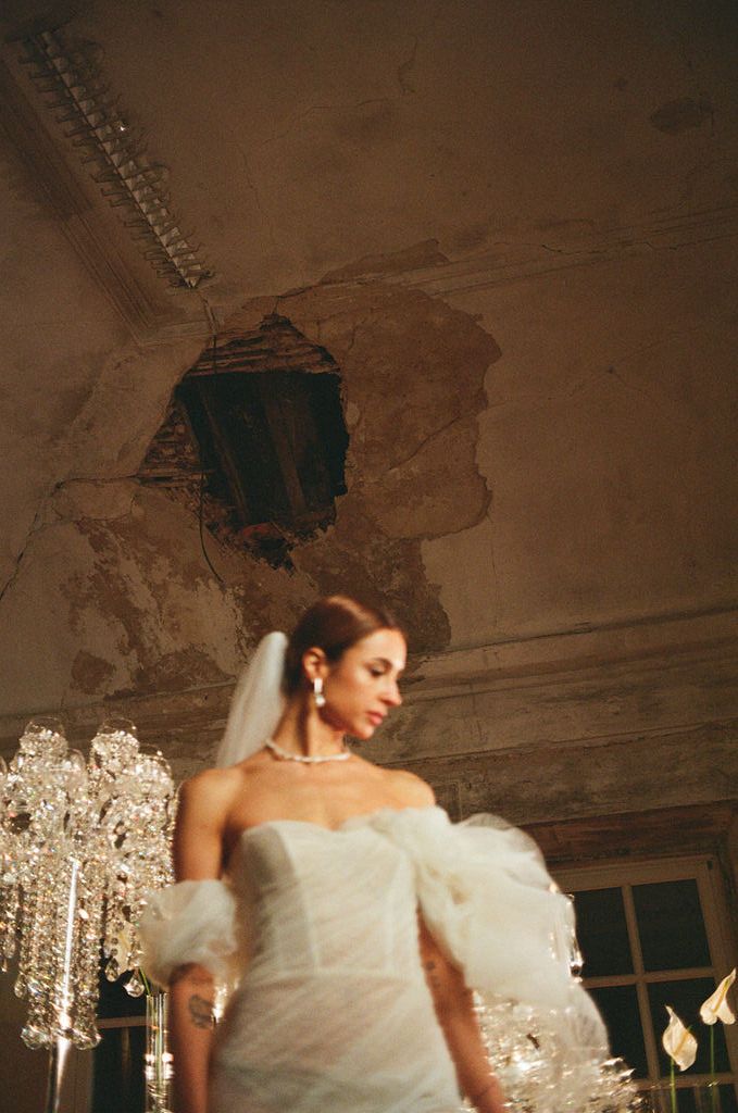 Bride in white off-shoulder gown with veil stands before crystal chandeliers in a rustic venue with damaged ceiling.
