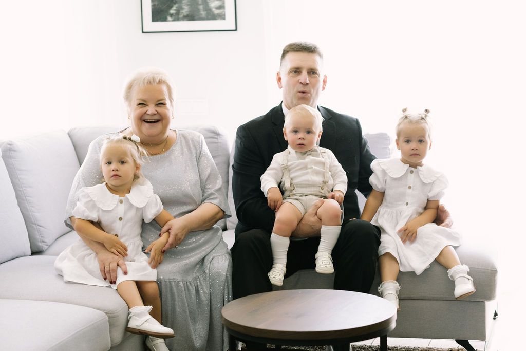 A happy family with grandmother, father, and three young children pose together on a gray sofa for a portrait.