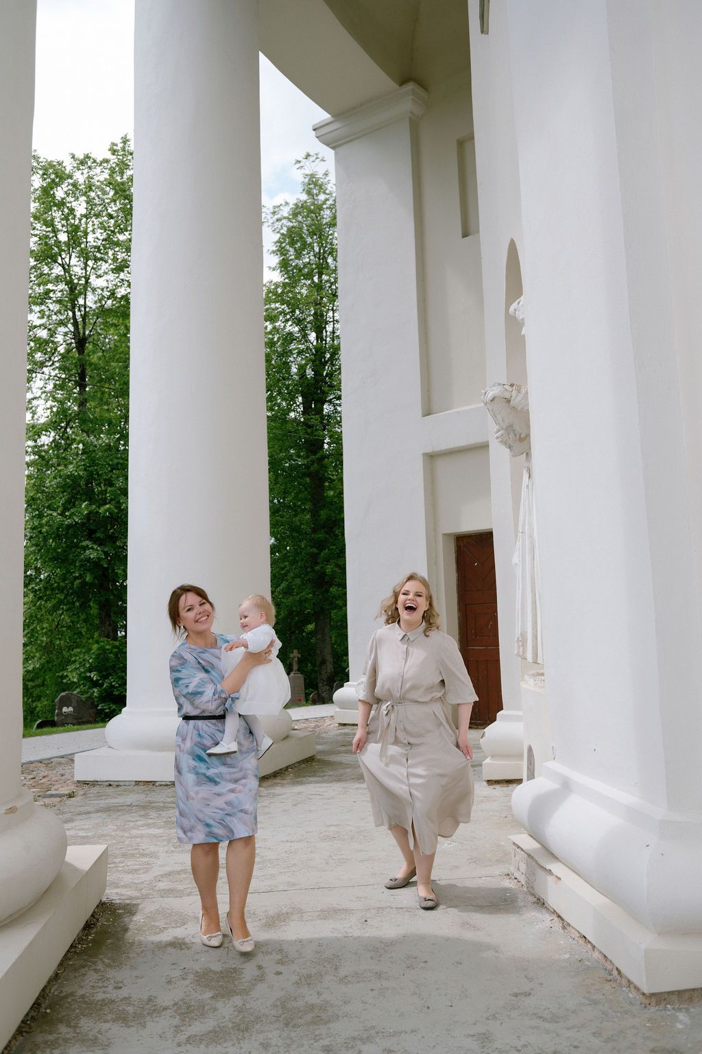Two women, one holding a baby, walk and laugh near white columns of a classical building outdoors.
