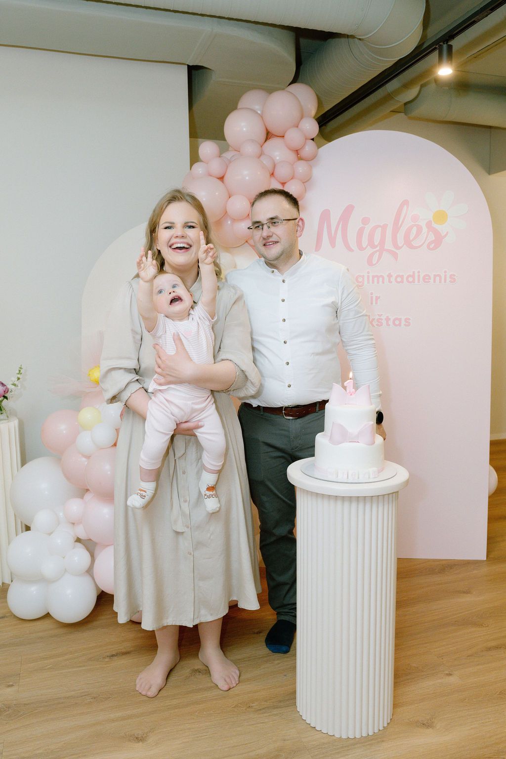 Family celebrating baby Migle's first birthday party with pink balloons and a white cake.