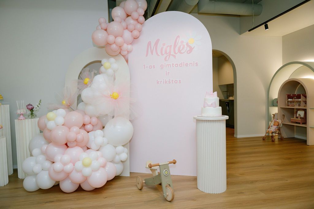 Pink balloon garland decorates a party backdrop with a white arch sign, cake display, and wooden toy bike.